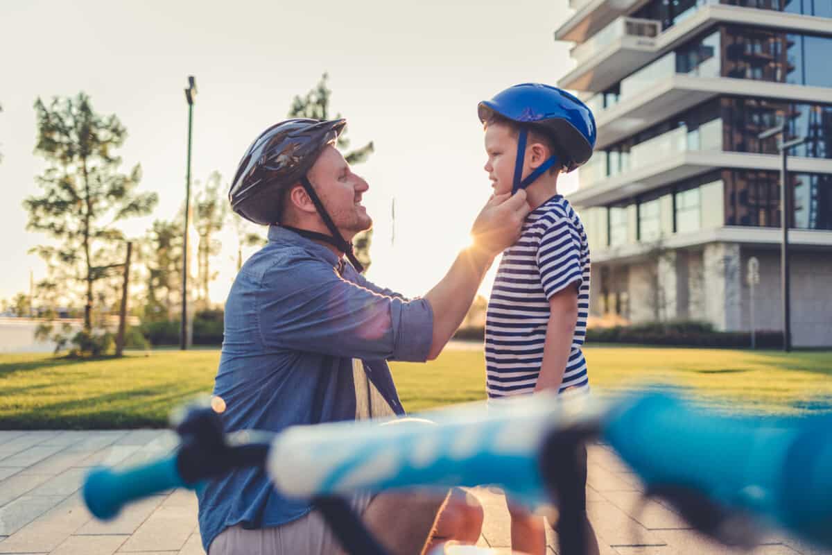 Father helping cheerful son wearing helmet for cycle. Excited little boy getting ready by wearing bike helmet to start cycling. Happy cute boy learn to ride a bike with his dad.
